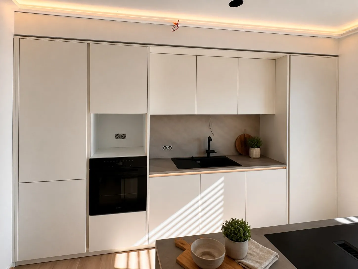 Grifflose Schrankwand in Cremeweiß mit eingebautem Backofen, schwarzer Spüle und Betonoptik-Rückwand